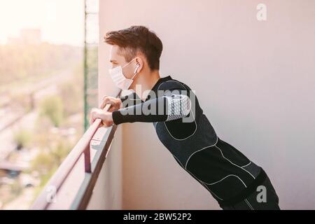 Sportlicher junger Mann Fitness-Trainer in medizinischen Gesichtsmaske tun Liegestützübungen auf dem Balkon. Selbstbewusste Sportmann in Schutzmaske Training worko Stockfoto