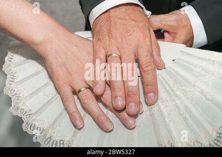 Hand des Bräutigams mit einem goldenen Ring auf der Hand der Braut auch mit einem Ring und beide Hände auf einem weißen Fächer. Stockfoto