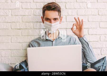 Junger Hipster Mann in schützender Gesichtsmaske zeigt okay Zeichen, während Online-Videoanruf zu Hause. Glücklicher Mann in medizinischen Gesichtsmaske sitzen auf Couch, sprechen Stockfoto