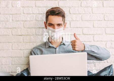 Glücklich Hipster Mann in medizinischen Gesichtsmaske sitzen auf Couch und Gesturing Daumen bis zu Laptop, im Web virtuellen Chat sprechen. Junger Mann in medizinischen Gesichtsmaske havi Stockfoto