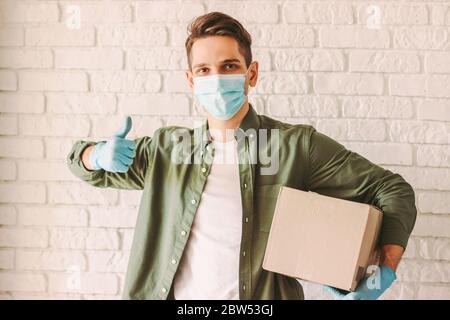 Glücklich Hipster Mann Kurier in medizinischen Gesichtsmaske zeigt Daumen bis Zeichen Geste und halten Karton in der Hand. Mann Lieferdienst Mitarbeiter trägt pro Stockfoto