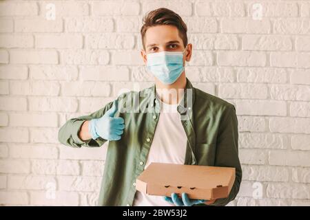 Glückliche Lieferung Mann in medizinischen Gesichtsmaske und Handschuhe halten Karton Pizza-Box in der Hand, zeigen Daumen bis Zeichen. Hipster Kerl Kurier in schützende Gesichtsmaske c Stockfoto