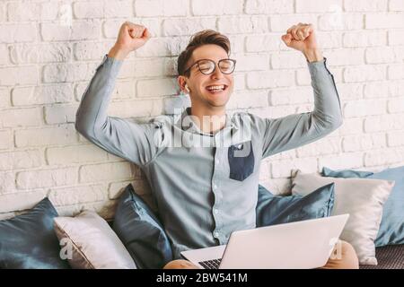 Glücklich Hipster Mann freiberuflicher Brillen jubeln Erfolg während der Arbeit zu Hause mit Laptop. Der junge Geschäftsmann ist fröhlich und lächelt. Emotional Stockfoto
