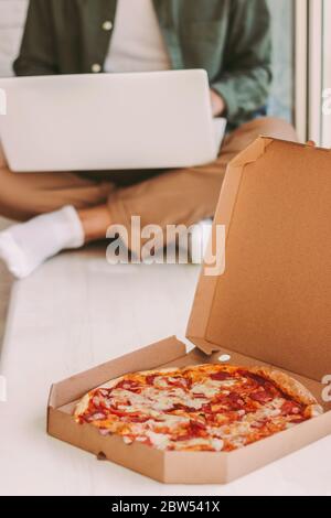 Nahaufnahme von Karton mit frischen leckeren Käse Pizza auf Holzfensterbank. Hipster Mann freiberuflicher Arbeit mit Laptop auf Hintergrund. Leckeres Iitali Stockfoto
