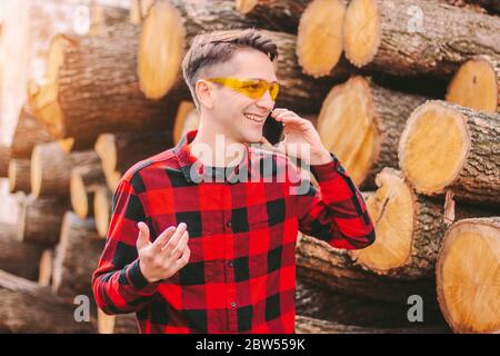 Junge glückliche Mann Sägewerk Lager Arbeiter in Schutzbrille sprechen Handy und lächelnd. Geschäftsmann Besitzer von kleinen Holzwerkstoff Produktion Stockfoto