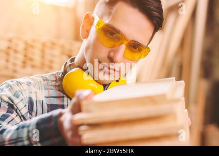 Nahaufnahme von selbstbewussten Schrankhersteller in Schutzbrille und Kopfhörer halten Stapel von Holzmaterial in den Händen. Junger Zimmermann, der bei SAW arbeitet Stockfoto