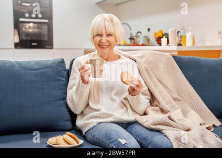 Ruhestand. Ältere Frau, die zu Hause mit Cookies sitzt und fröhlich lacht Stockfoto