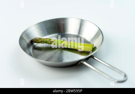 Konzeptfoto, riesiger Spargel auf einer kleinen Stahlpfanne auf weißem Hintergrund. Stockfoto