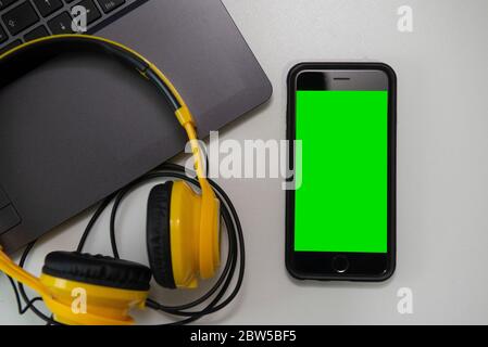 Ansicht von oben auf den Schreibtisch mit offenem Laptop, Kopfhörer und Smartphone mit grünem Bildschirm. Nahaufnahme, um das Bild auf dem Bildschirm des Mobiltelefons einzufügen. Selektiver Fokus Stockfoto