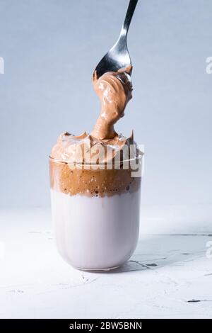 Oreischer Dalgona Kaffee und Löffel mit Sahne Kaffee fällt in ein Glas auf weißem Hintergrund. Vertikales Bild. Stockfoto
