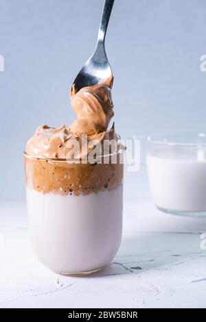 Dalgona Kaffee und Schlagkaffee fallen aus einem Löffel in ein Glas. Koreanischer trendy, flauschig, cremig und Schlagsahne Eistkaffee. Vertikales Bild. Stockfoto