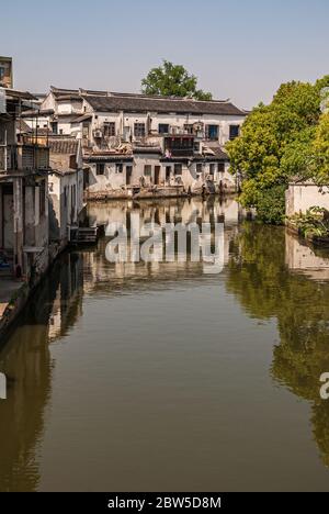 Tongli, Jiangsu, China - 3. Mai 2010: Linie von dunkel überdachten weißen Häusern, die sich im braunen Wasser des Kanals unter hellblauem Himmel spiegeln. Grünes Laub und rotes l Stockfoto