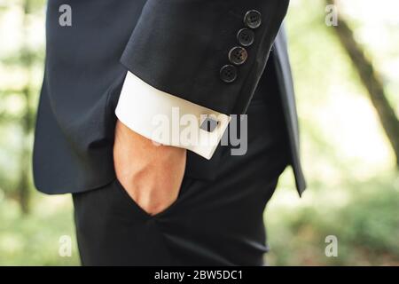 Seitenansicht von Männern in schwarzem Anzug. Nahaufnahme der Hand in der Tasche. Schwarze quadratische Manschettenknöpfe an weißen Hemdärmeln. Hintergrund im Freien. Stockfoto