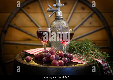 Horizontale Fine Art Bild von zwei Gläsern Rotwein sitzen auf einem Holzfass mit Trauben und einem verschwommenen Wagenrad im Hintergrund geschmückt. Stockfoto