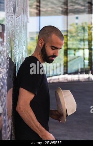 Junger Mann steht gegen silberne Wand mit einem Hut in der Hand Stockfoto