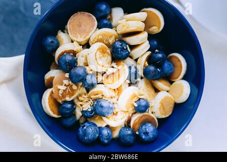 Nahaufnahme von Mini Pancake Müsli, Mini Pfannkuchen in einer dunkelblauen Schüssel mit Ahornsirup Honig mit Heidelbeere. Hintergrund der Speisen. Stockfoto