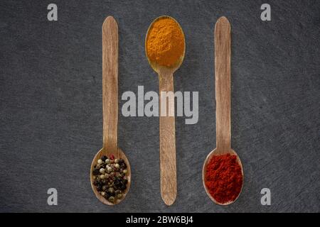 Drei Holzlöffel mit farbigem Pfeffer, gemahlener Kurkuma und Rosenpeppers auf einem dunklen Schieferteller Stockfoto