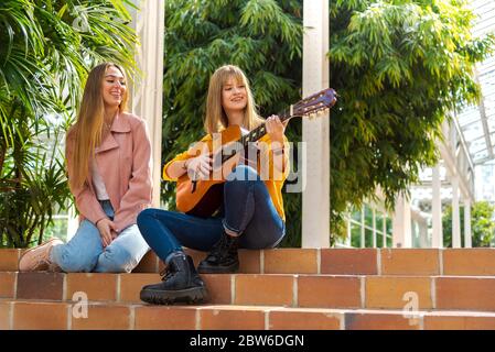 Zwei attraktive junge kaukasische Mädchen mit blonden Haaren lächeln glücklich, während man eine Gitarre spielt, die auf einer Treppe mit Bäumen im Hintergrund sitzt Stockfoto