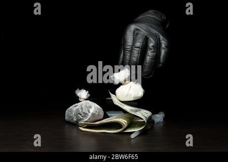 Eine Hand mit schwarzen Handschuhen nimmt vom Tisch oder legt eine Drogentasche darauf. Andere Drogen und Geld sind bereits auf dem Tisch. Stockfoto