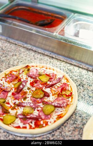 Rohe Pizza mit Wurst, Käse, Gurken und Tomatensauce zum Backen in der Küche bereit. Stockfoto