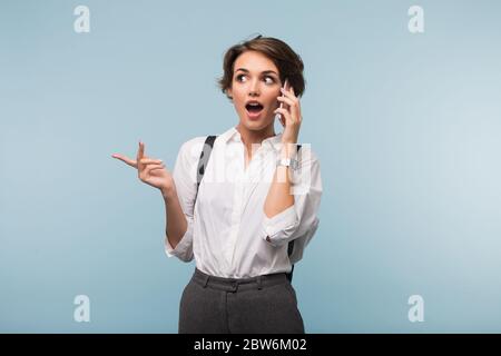 Junge Frau mit dunklen kurzen Haaren in weißem Hemd, die beim Telefonieren auf blauem Hintergrund verblüfft zur Seite schaut Stockfoto