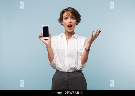 Junge, staunende Dame mit dunklen, kurzen Haaren in weißem Hemd, die das Handy in der Hand hält, mit offenem Mund, der in der Kamera auf blauem Hintergrund schaut Stockfoto