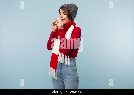 Junge ungesunde Dame mit dunklen kurzen Haaren in roten Pullover, Schal und Strickmütze Niesen mit weißen Servietten in Händen über blauen Hintergrund isoliert Stockfoto