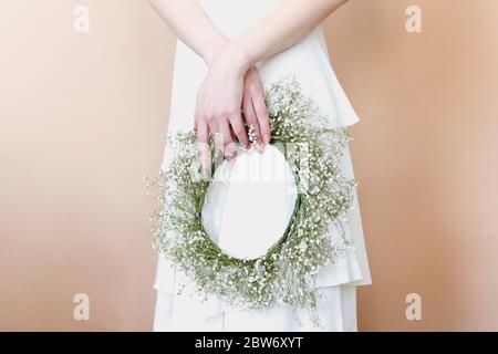 Ernte anonyme junge Frau in weißem Brautkleid mit zarten Kranz mit weißen Blumen gemacht, während sie vor beigem Hintergrund stehen Stockfoto