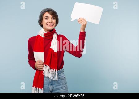 Ziemlich lächelndes Mädchen mit dunklen kurzen Haaren in roten Pullover, Schal und Strickmütze glücklich in der Kamera suchen, während das Halten weißen Papier Form der Nachricht in Stockfoto