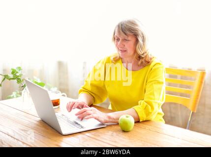 Positive Frau mittleren Alters in Freizeitkleidung sitzen am Holztisch mit einer Tasse Tee und Apfel und Surfen Laptop während der Morgenzeit zu Hause Stockfoto