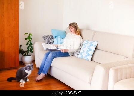 Ganzkörper-Seitenansicht von ruhigen barfuß mittleren Alters Frau in lässigen Kleidung sitzen auf dem Sofa und lesen interessante Zeitschrift während der Ruhe in gemütlichen l Stockfoto