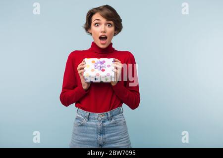 Schöne überrascht Mädchen mit dunklen kurzen Haaren in roten Pullover erstaunlich in der Kamera zu suchen, während kleine Geschenk-Box in Händen über blauem Hintergrund Stockfoto