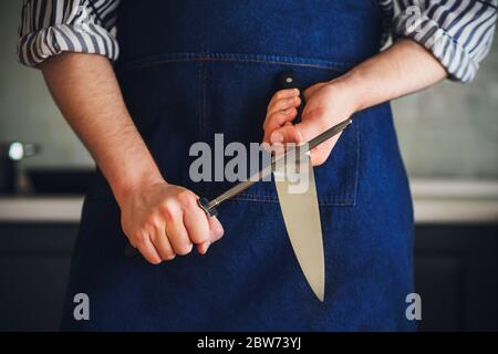 Ein Mann, der in eine blaue Küchenschürze und ein gestreiftes Hemd gekleidet ist, schärft die Klinge eines Messers mit einem Wetzstein, bevor er mit dem Kochen beginnt. Hausmannskost. Küche Stockfoto