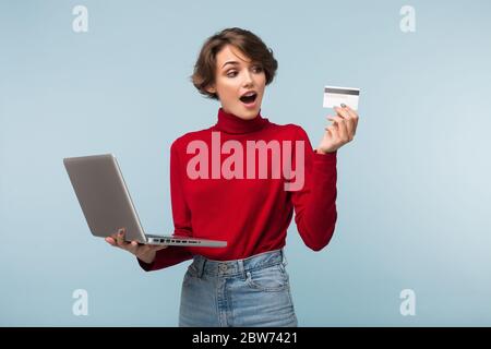 Junge Frau mit dunklen kurzen Haaren in rotem Pullover und Jeans, die Laptop in der Hand hält, während sie erstaunt auf Kreditkarte über blauem Hintergrund schaut Stockfoto