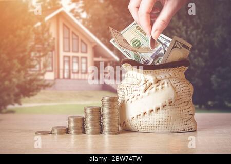 Hand setzen Münze in Tasche mit US-Dollar-Rechnungen, Münzen Stapel auf Tisch, verschwimmen Hintergrund mit Haus. Konzept der Geldeinsparung. Anlageideen, Finanzmanaa Stockfoto