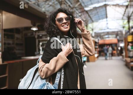 Cool afroamerikanischen Mädchen trägt Sonnenbrille und Kopfhörer. Lächelndes Mädchen mit dunklen lockigen Haaren suchen in der Kamera auf der Straße schönes Mädchen im Mantel, Schal und Stockfoto
