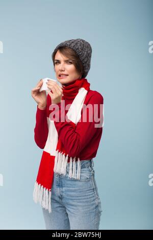 Junge ungesunde Frau mit dunklen kurzen Haaren in roten Pullover, Schal und Strickmütze, die weiße Serviette in den Händen hält, während frustriert in der Kamera zu suchen Stockfoto