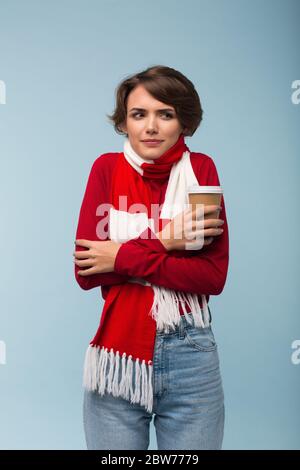 Junge Frau in roten Pullover und Schal hält Tasse Kaffee in der Hand nachdenklich beiseite schauen, während Schütteln von Kälte über blauem Hintergrund gehen Stockfoto