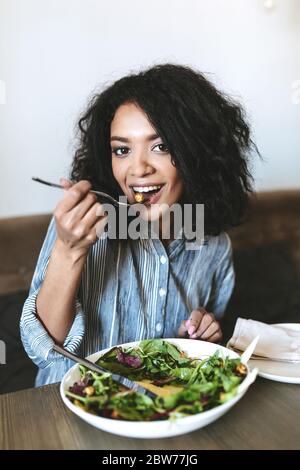 Nettes afroamerikanisches Mädchen, das Salat im Restaurant isst. Porträt von lächelnden Dame mit dunklen lockigen Haaren und Salat auf dem Tisch im Café schöne Mädchen mit Stockfoto