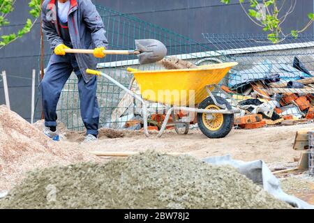 Ein Arbeiter in einem dunkelblauen Overall mit einer Schaufel lädt Sand in einer gelben Baukarre vor dem Hintergrund einer Baustelle. Stockfoto