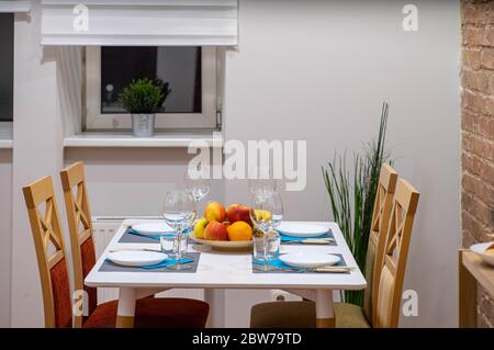 Nahaufnahme des Tisches mit Holzstühlen. Teller und Weingläser. Fenster mit Jalousien. Moderne Dachgeschosseinrichtung. Ziegelwand. Stockfoto