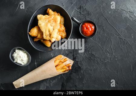 Fisch und Chips in einem Papier-Kegel-Dip und Zitrone - gebratener Kabeljau, pommes frites, Zitronenscheiben, Tartarsauce, Ketchup Tomatoe und matschige Erbsen über schwarzem Backgrou Stockfoto