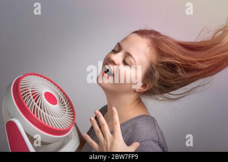 Junge kaukasische Frau Mädchen mit langen Haaren hält elektrischen Ventilator vor ihrem Gesicht. Kühlen Sie sich ab bei heißem Sommerwetter Stockfoto