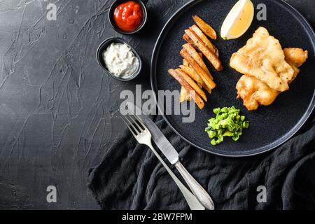 Fischchips mit Dip und Zitrone - gebratener Kabeljau, pommes frites, Zitronenscheiben, Tartarsauce, Ketchup-Tomate und matschige Erbsen mit Gabel und Messer auf schwarzem Teller Stockfoto