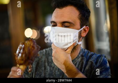 Junger Mann versucht, ein Schweinefleisch Shank mit einer Maske, lustige Coronavirus-Konzept zu essen Stockfoto