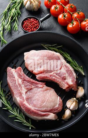 Rohes Schweinefleisch Lenden Koteletts in Pfanne in der Nähe Zutaten wtih Kräuter Pfeffer und Tomaten Draufsicht vertikale selektive Fokus noone. Stockfoto