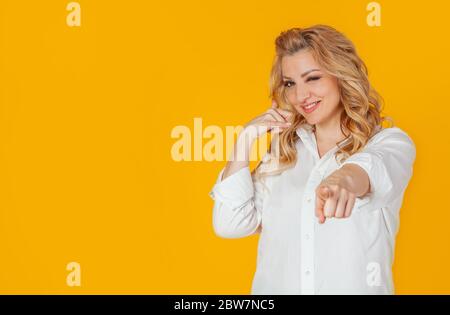 Porträt einer schönen blonden Frau in einem weißen Hemd, zeigt auf die Kamera, lächelt und macht eine Telefongeste, als ob sie nach einer Nummer fragt, mit jemandem flirtet, auf einem gelben Hintergrund steht. Stockfoto