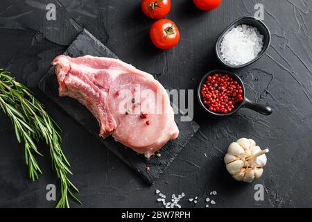 Draufsicht rohes Schweinekotelett Steak und Salz, Pfeffer auf schwarzem Schiefer Stein Brett mit Rosmarin und frische Zutaten für Grill über schwarzem strukturierten Hintergrund Stockfoto