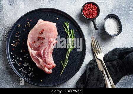 Rohe Schweineloin Koteletts in einem schwarzen runden Teller auf einem grauen strukturierten Steinhintergrund mit Rosmarin Knoblauch Pfefferkörner Zutaten für Grill Draufsicht. Stockfoto