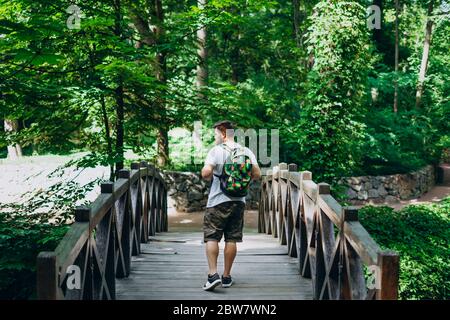 Sofia Park, Uman. Schöner Mann mit Rucksack auf einer Holzbrücke. Männlicher Wanderer auf einer Brücke zwischen grünen Sommerpark. Spaziergang im Sommer grün f Stockfoto